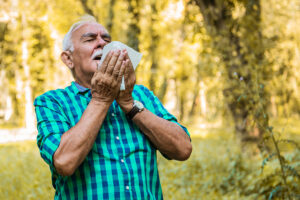 Older man standing in a wooded area and struggling with allergy symptoms, holding up a tissue as he prepares to sneeze