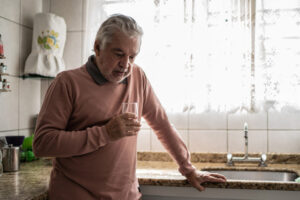 An older man stands in the kitchen holding a glass while thinking about the importance of managing depression in cardiac recovery.